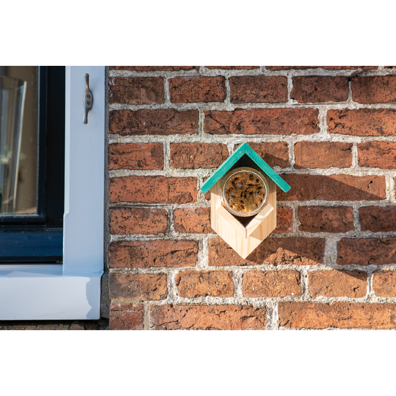 A wooden bird feeder mounted on a brick wall, holding a peanut butter jar, with a green slanted roof.